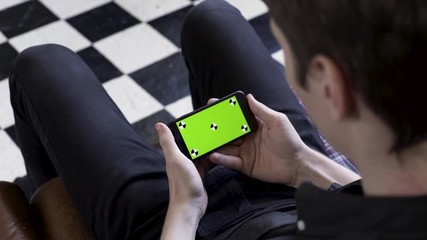 Poster - Business man using smart phone with green screen while sitting on a couch on checkered black and white floor background. Stock footage. Man holding device in horizontal position and watching.