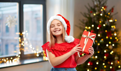 Poster - childhood and winter holidays concept - smiling happy girl in santa helper hat with gift box over christmas tree at home background