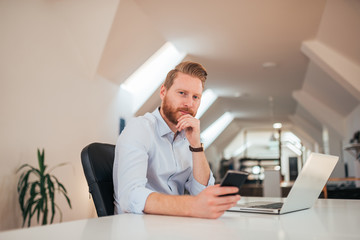 Wall Mural - Portait of young redhead entrepreneur using smartphone and laptop at work.