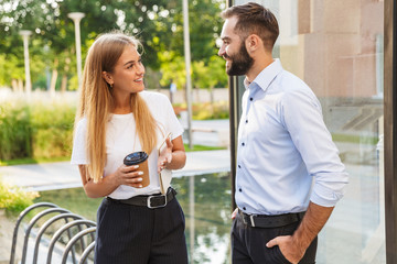 Smiling man and woman businesspeople outside at the street near business center holding cup of coffee talking with each other.