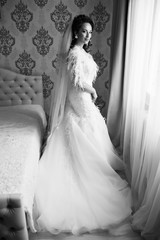 Bride morning preparation. Beautiful bride in white wedding dress and veil near  bedroom window. Adorable fiancee waiting for the groom. Young brunette bride in the morning at home. Black and white