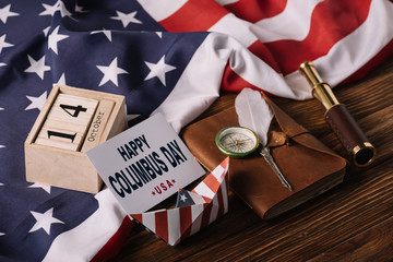 Sticker - card with happy Columbus Day inscription near calendar, paper boat, nib, compass, telescope and notebook on wooden surface with American national flag