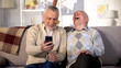© motortion - Elderly man showing funny photo on smartphone aged friend, having fun at home