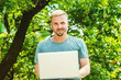 © Alexander Image - Young Happy American Man with beard, working in New York City, wearing light green T shirt, sitting outside with green trees in school campus in summer, working on laptop computer, looking, smiling..