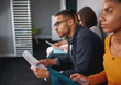 © StratfordProductions - Side view of a confident young african american man wearing eyeglasses holding resume in hand waiting for job interview with other candidates