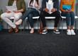 © StratfordProductions - Low section of job applicants waiting in corridor preparing for recruiting process - photo of diverse feet sitting in a row waiting for job interview