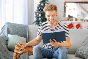 Poster - Young man with cute cat reading book at home on Christmas eve