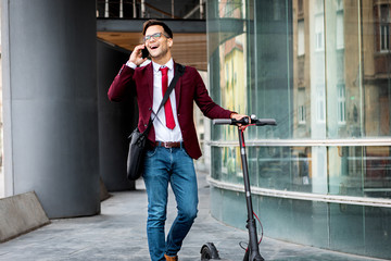 Wall Mural - young businessman with electric scooter standing in front of modern business building talking at pho