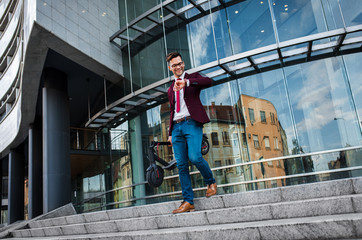 Wall Mural - Young businessman with electric scooter standing in front of modern business building looking at watch.