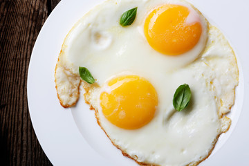 Two fried eggs in a white ceramic plate with basil leaves on an old wooden table. Dark background.