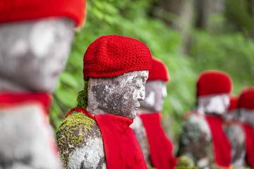 Close up of a row of stone japanese jizo statues wearing red caps and bibs, against a green bokeh background