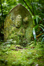 Close Up Of A Low, Ancient And Slanting Japanese Tombstone Covered With Moss, Lying Among Green Vegetation And Engraved With The Basrelief Of A Buddha