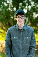 Wall Mural - Teenage boy with glasses standing outside in front of a tree wearing a gray button up long sleeved shirt and denim jeans