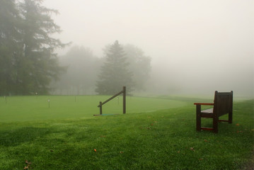 Wall Mural - early autumn foggy morning landscape. scenic view with deep fog over a golf course with wooden bench