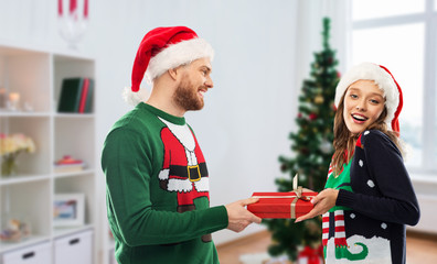 Poster - people and winter holidays concept - happy couple in santa hats with gift in ugly sweaters over christmas tree at home background