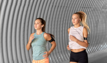 fitness, sport and healthy lifestyle concept - young women or female friends with earphones wearing armbands with smartphones and running outdoors