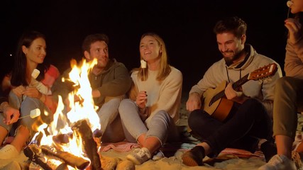 Poster - leisure and people concept - group of smiling friends sitting at camp fire on beach, roasting marshmallow and playing guitar at night