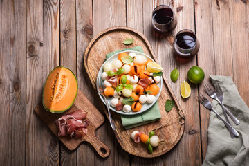Melon, mozzarella, prosciutto salad, appetizer or snack, two glasses of red wine, party food, top view, wooden background