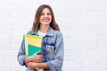 Girl Student Holds Folders And A Notebook In Her Hands And Smiles On A Background Of A White Brick Wall, Copy Space