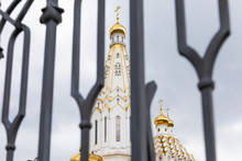 White Christian Church With Gilded Domes Seen Through Defocused Black Wrought Fence
