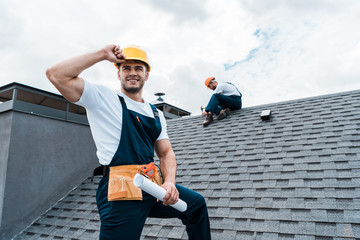 Wall Mural - selective focus of happy repairman holding rolled paper while coworker repairing roof