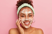Teenage Skincare Concept. Optimistic Dark Skinned Lady Uses Foaming Cleanser For Washing Face, Smiles Gently, Stands Naked, Shows Bare Shoulders, Looks Straightly At Camera Poses Over Rosy Studio Wall