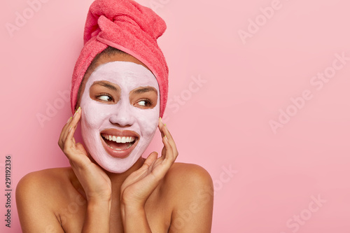 spa-girl-with-healthy-fresh-dark-skin-applies-nourishing-clay-mask-stands-with-bare-shoulders-looks-happily-away-has-wrapped-towel-on-head-takes-bath-looks-aside-with-cheerful-expression
