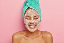 Headshot Of Pretty Smiling Woman Applies Salt Granules On Face, Keeps Eyes Closed, Shows White Perfect Teeth, Wears Turquoise Towel, Poses Shirtless Against Pink Background, Enjoys Effective Result