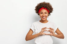 Love To You, My Dear. Glad Satisfied African American Woman Makes Heart Gesture, Smiles Pleasantly At Camera, Wears Casual Outfit, Expresses Good Feelings To Lover, Poses Indoor Over White Background