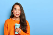 Horizontal Shot Of Pretty Brunette Japanese Woman Has Long Hair, Wears Vivid Orange Warm Sweater, Holds Coffee To Go, Imagines Pleasant Moment In Life During Break, Models Over Blue Studio Wall