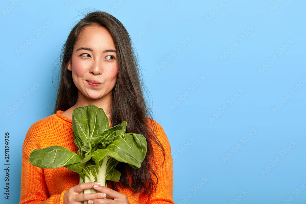 Photo of attractive young woman holds fresh green vegetable, eats healthy food at home, uses food product for making vegetarian salad, wears orange jumper, poses indoor. Home growing concept. - obrazy, fototapety, plakaty 