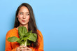 © wayhome.studio  - Photo of attractive young woman holds fresh green vegetable, eats healthy food at home, uses food product for making vegetarian salad, wears orange jumper, poses indoor. Home growing concept.