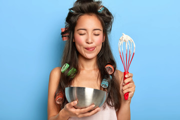 Brunette lady licks tongue, makes butter cream, holds bowl and hand whisk, beats egg white, wears curlers on dark long hair, nightdress, busy cooking something delicious at kitchen for husband