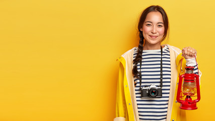 Wall Mural - Studio shot of pleased brunette woman dressed in raincoat, striped jumper, carries retro camera and oil lamp, enjoys adventurous journey, has long pigtail, isolated over yellow wall, copy space aside