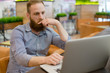 © ruslanshug - pensive young caucasian man with beard sitting with laptop indoor cafe solving problems online wi fi
