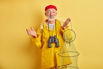 Wall Mural - puzzled confused bearded old man with binocular and fishnet in hands shrugging his shoulders, close up photo. unsuccessful day for fishing. close up photo