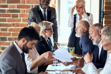 Wall Mural - Group of businessmen wear suits sit at table in office and discuss new project, smile. caucasian blond woman and african man stand, behind open window. Camera focused on blond woman listen