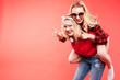 © Вячеслав Косько - Stock studio portrait of two cheerful and positive trendy girlfriends or sisters having fun. Girl in sunglasses making bunny ears to her friend with her hand while sitting on her back. Isolate on red.