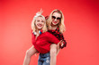 © Вячеслав Косько - Stock studio portrait of two cheerful and positive trendy girlfriends or sisters having fun. Girl in sunglasses making bunny ears to her friend with her hand while sitting on her back. Isolate on red.