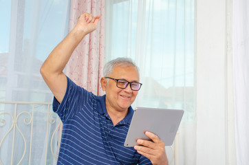 Senior man sitting on sofa are feeling glad and delightful when looking at tablet. Senior man raising the hand and yelling out , Happy retirement concept.