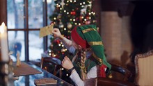 Cute Elf With Envelope On The Background Of Christmas Lights. Happy Female Elf Looking At Envelope Attentively While Sitting At The Table.