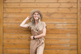 Fototapeta Na drzwi - Portrait of a smiling attractive caucasian blonde woman in stylish outfit and hat posing on a wooden background. Charming model posing in summer clothes. Advertising space