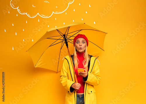 Fotografía Girl with smartphone and umbrella on yellow background surprised for the weather