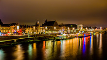 Night Cityscape Of The River Maas With Its Promenade, Pier With A Boat Anchored With Buildings, Illuminated By Multi-colored Lights And Reflected On The Water, Maastricht, South Limburg, Netherlands