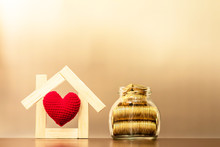 Red Heart In The Middle Of The Wooden House Model And Gold Coin In The Bottle Bank With Growing Interest On Brown Background, Business Investment Or Saving Money To Buy Real Estate For Family Concept.