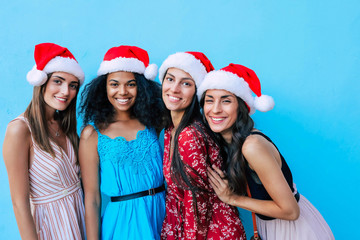 Wall Mural - Before the party. A group of four beautiful girls in Santa hats standing in a row hugging and looking into the camera over turquoise background