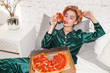 young fashionable woman in silk pajamas lays on bed and eat pizza pepperoni from pizza box, direct flash