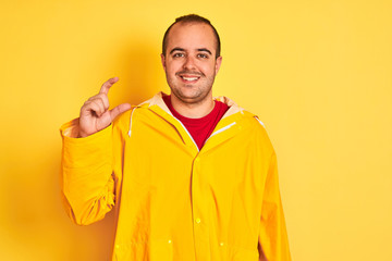 Sticker - Young man wearing rain coat standing over isolated yellow background smiling and confident gesturing with hand doing small size sign with fingers looking and the camera. Measure concept.