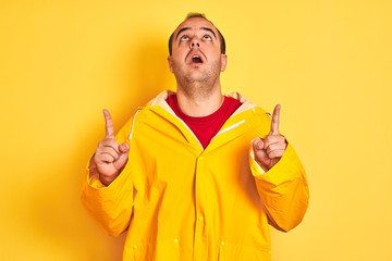 Sticker - Young man wearing rain coat standing over isolated yellow background amazed and surprised looking up and pointing with fingers and raised arms.