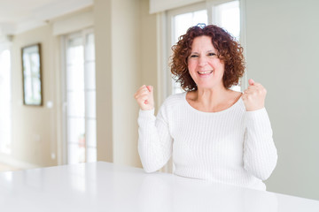 Poster - Beautiful senior woman wearing white sweater at home celebrating surprised and amazed for success with arms raised and open eyes. Winner concept.